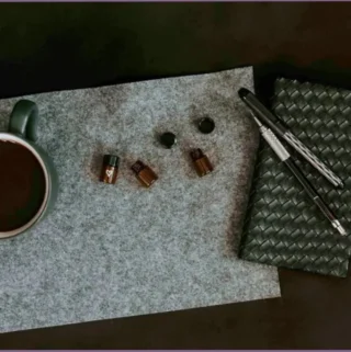 essential oil bottles and cup of coffee lying on grey felt mat with black binder and 2 pens lying at the side