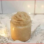 Christmas cookie sugar scrub in a glass jar, sitting on a white and silver snowflake towel with soft holiday lights and a metal ornament in the background