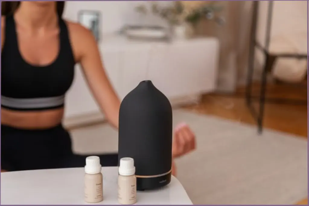 Aromatherapy diffuser releasing mist beside two essential oil bottles while a person sits cross‑legged in meditation - best essential oils for meditation