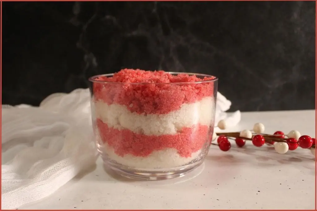 Alternating layers of red and white DIY candy cane sugar scrub in glass jar with a sprig of red berries in the background with FAQs on the recipe