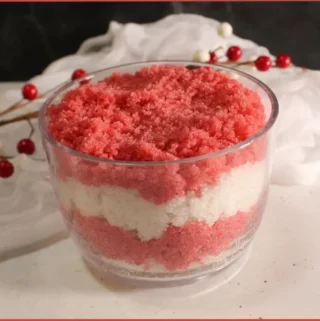 Alternating layers of red and white DIY candy cane sugar scrub in glass jar with a sprig of red berries in the background