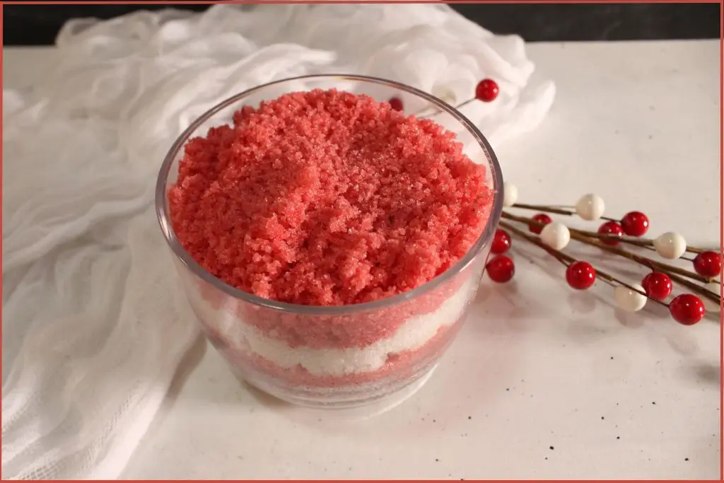Alternating layers of red and white homemade candy cane sugar scrub in glass jar with a sprig of red berries in the background with storage tips