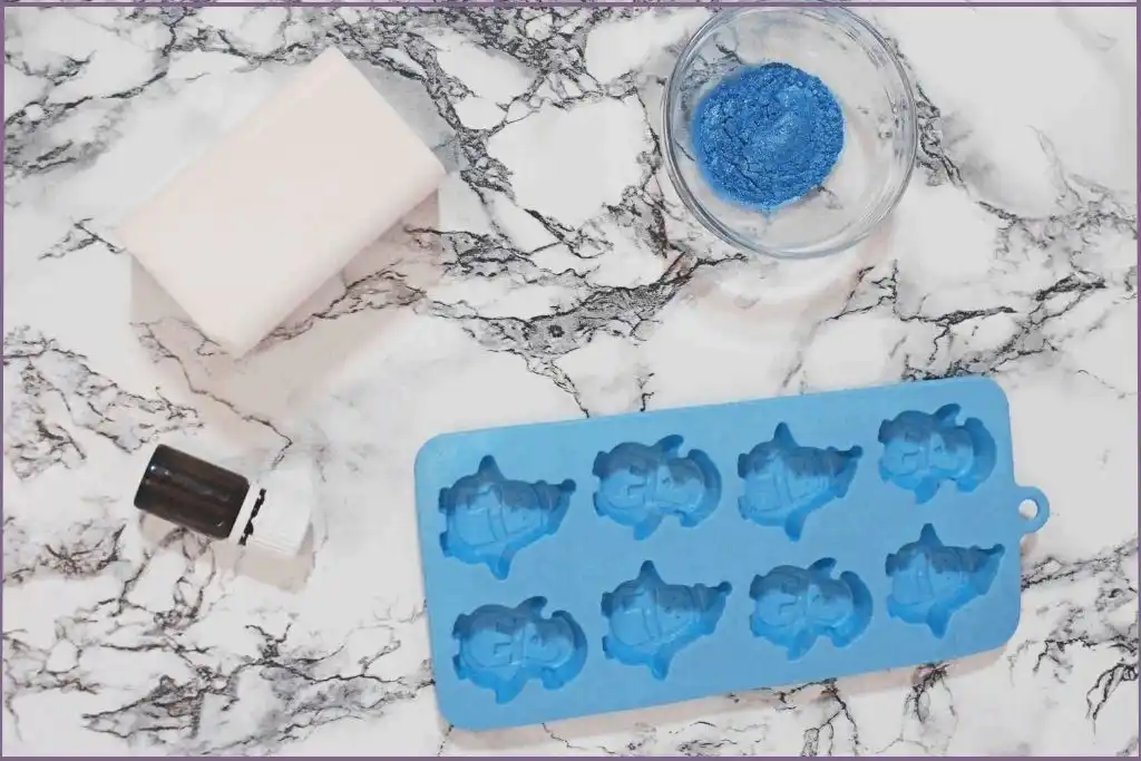 Ingredients for penguin soap recipe - penguin shaped soap mold, block of soap base, blue mica powder in a bowl and essential oil bottle