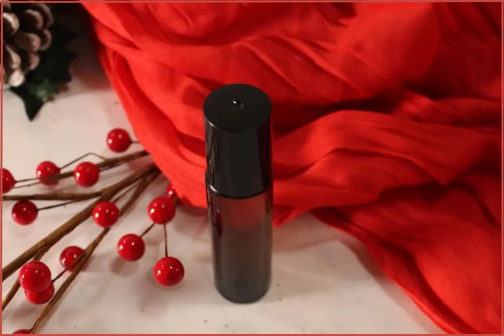Homemade Christmas roll-on perfume in a small amber bottle, styled with red berries, frosted pinecones, and a red scarf backdrop with ideas for gifting