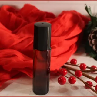 DIY Christmas roll-on perfume in a small amber bottle, styled with red berries, frosted pinecones, and a red scarf backdrop