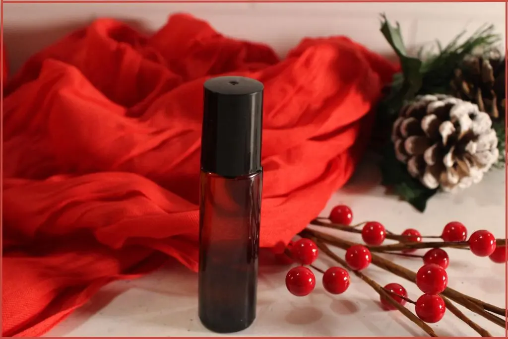 DIY Christmas roll-on perfume in a small amber bottle, styled with red berries, frosted pinecones, and a red scarf backdrop