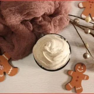 DIY gingerbread whipped body butter in a glass jar, creamy swirls on top, surrounded by gingerbread men decorations, soft pink cloth, and holiday pearl stems on a white background.