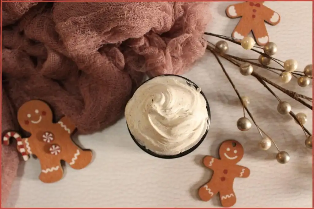 DIY gingerbread whipped body butter in a glass jar, creamy swirls on top, surrounded by gingerbread men decorations, soft pink cloth, and holiday pearl stems on a white background.
