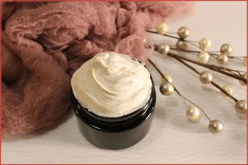 DIY gingerbread whipped body butter in a glass jar, creamy swirls on top, surrounded by soft pink cloth, and holiday pearl stems on a white background.