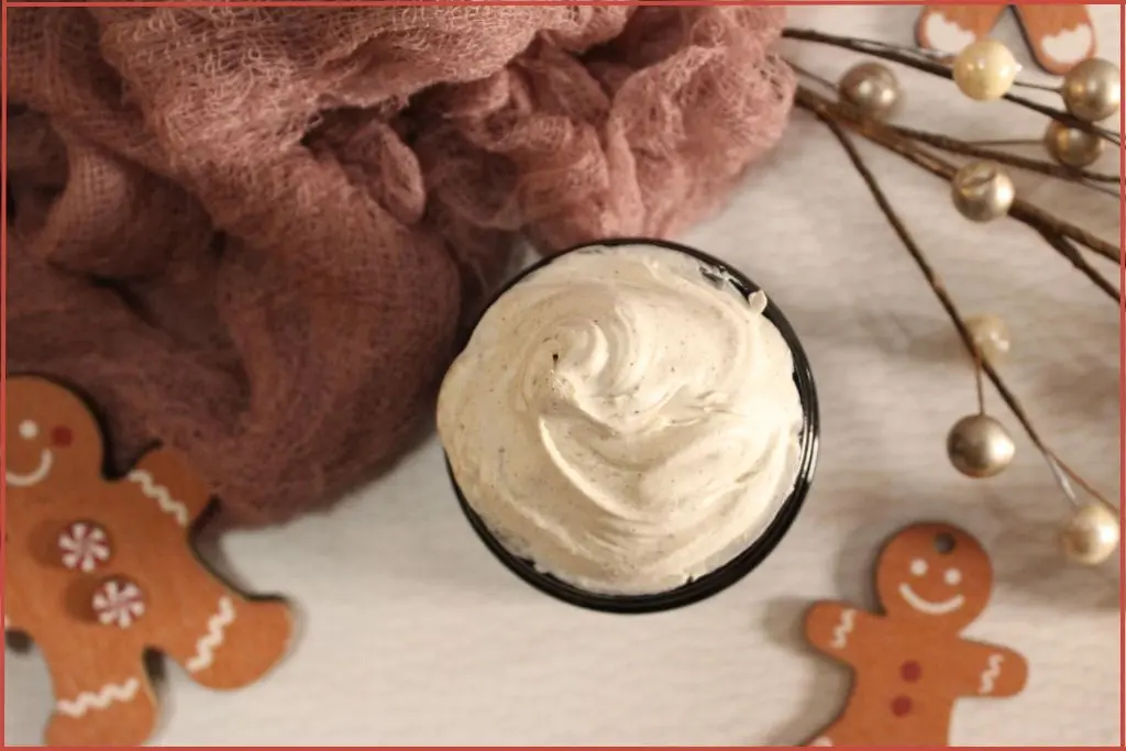 DIY gingerbread whipped body butter in a glass jar, creamy swirls on top, surrounded by gingerbread men decorations, soft pink cloth, and holiday pearl stems on a white background.