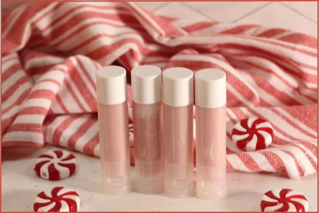 Four DIY candy cane lip balm tubes in front of a red‑and‑white striped towel, with peppermint candies scattered around.