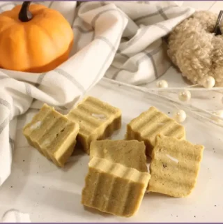 6 Rectangular homemade pumpkin spice sugar scrub bars with ridged tops arranged on a white surface, styled with a plaid kitchen towel, small orange pumpkin, and cozy pumpkin decor with instructions for DIY pumpkin spice sugar scrub bars.