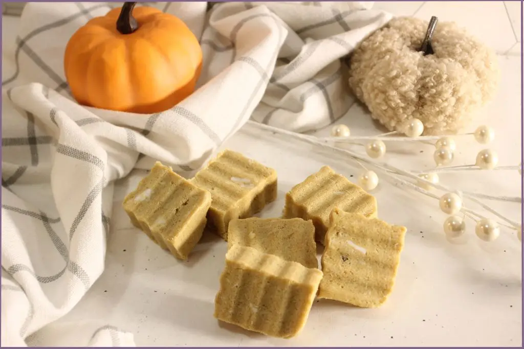 6 Rectangular homemade pumpkin spice sugar scrub bars with ridged tops arranged on a white surface, styled with a plaid kitchen towel, small orange pumpkin, and cozy pumpkin decor with instructions for DIY pumpkin spice sugar scrub bars.