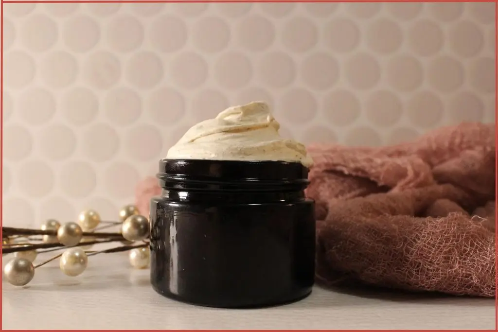 DIY gingerbread whipped body butter in a glass jar, creamy swirls on top, surrounded by soft pink cloth, and holiday pearl stems on a white background.