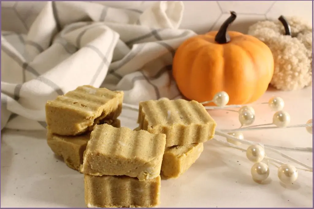 6 Rectangular homemade pumpkin spice sugar scrub bars with ridged tops arranged on a white surface, styled with a plaid kitchen towel, small orange pumpkin, and cozy pumpkin decor with instructions for how to use homemade pumpkin spice sugar scrub bars.