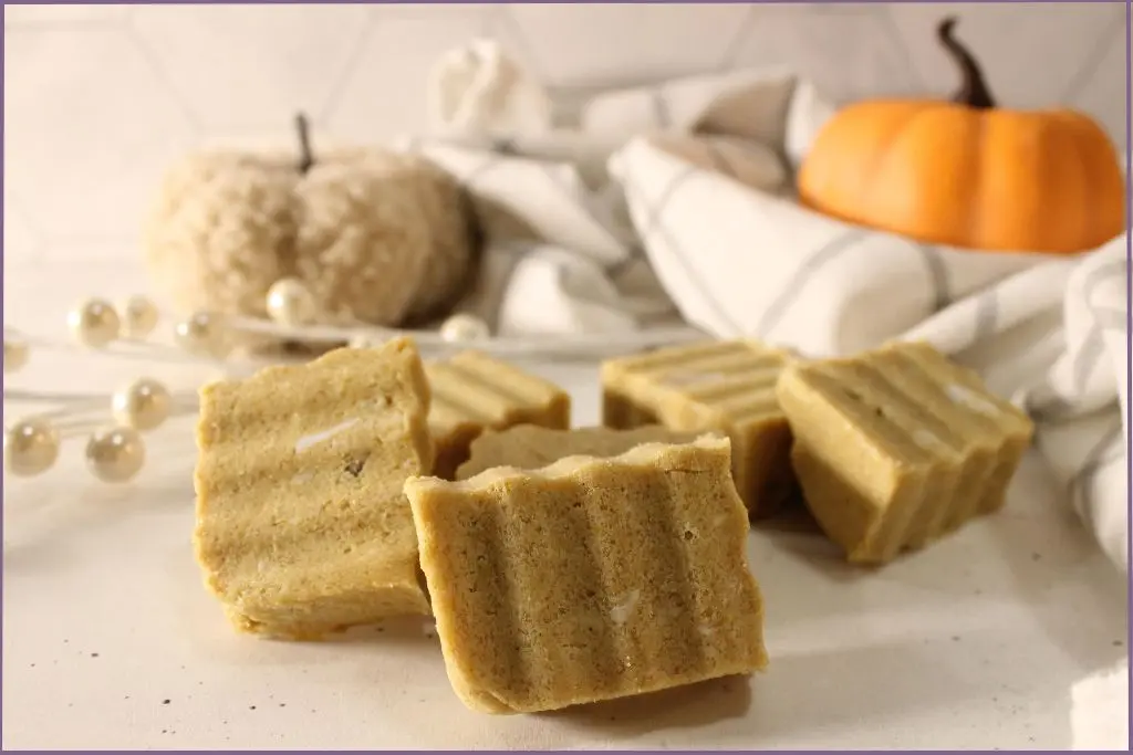 6 Rectangular homemade pumpkin spice sugar scrub bars with ridged tops arranged on a white surface, styled with a plaid kitchen towel, small orange pumpkin, and cozy pumpkin decor for a fall DIY beauty post.