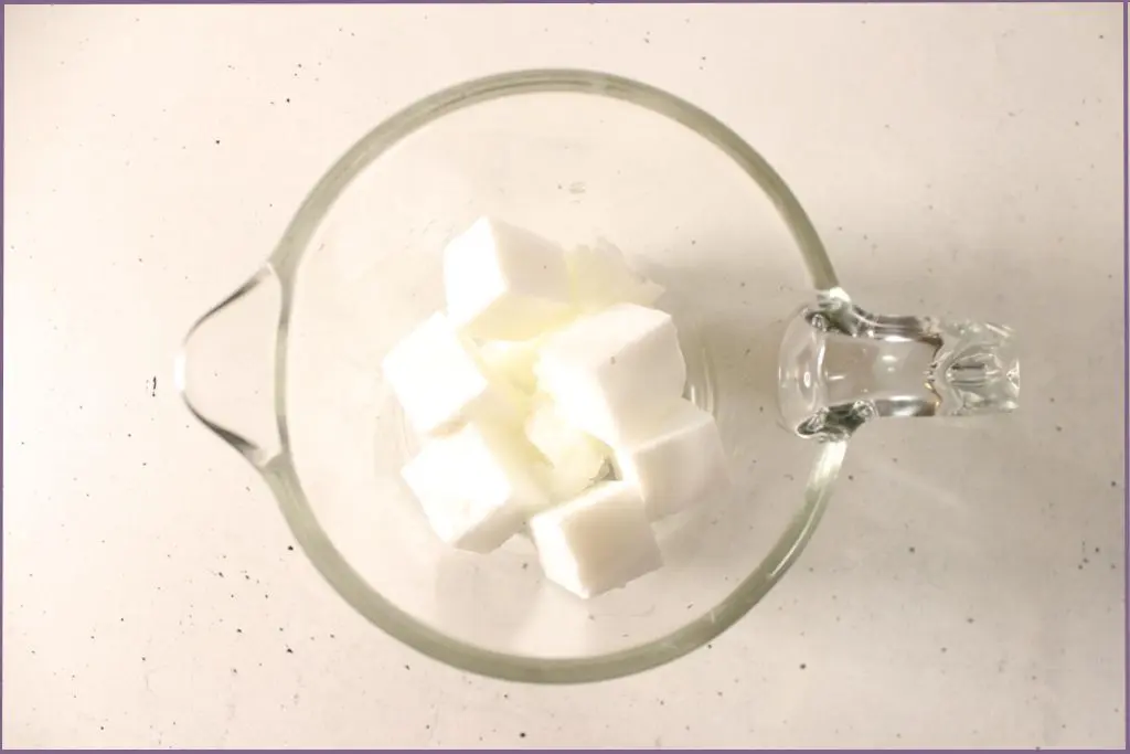 Glass measuring cup with small cubes of melt and pour soap base before melting on a slow cooker