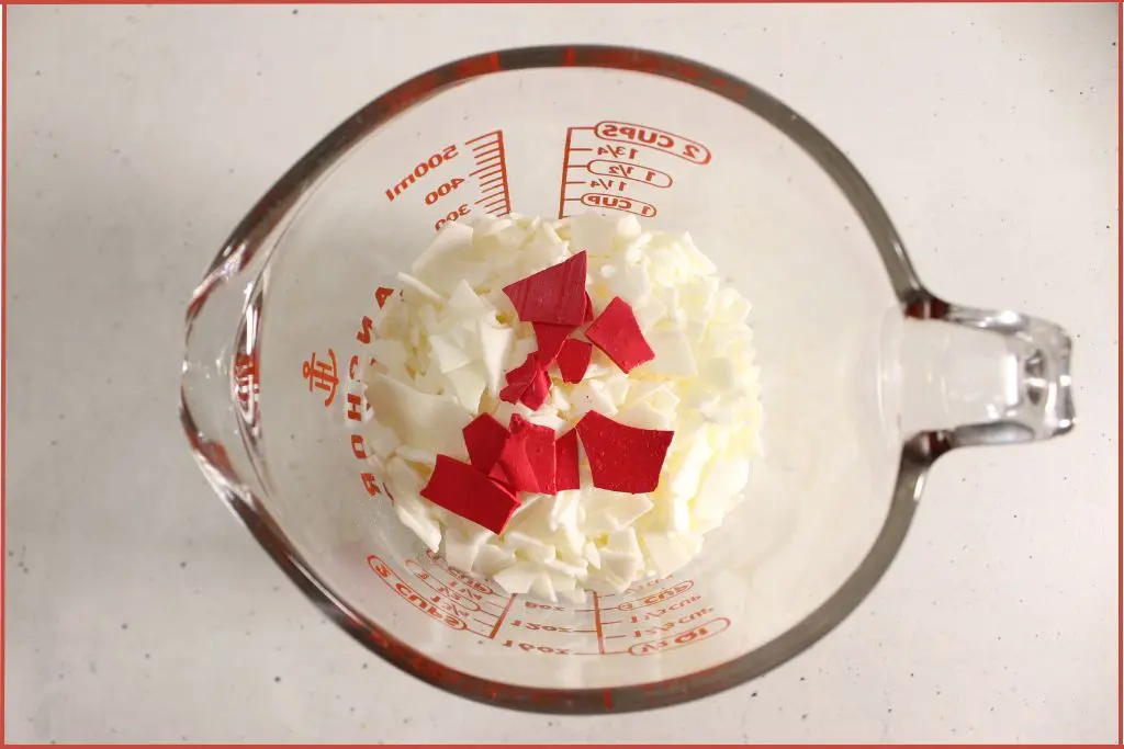 Overhead view of white soy wax flakes and red candle dye in a glass heatproof jar - prep to make a candy cane candle