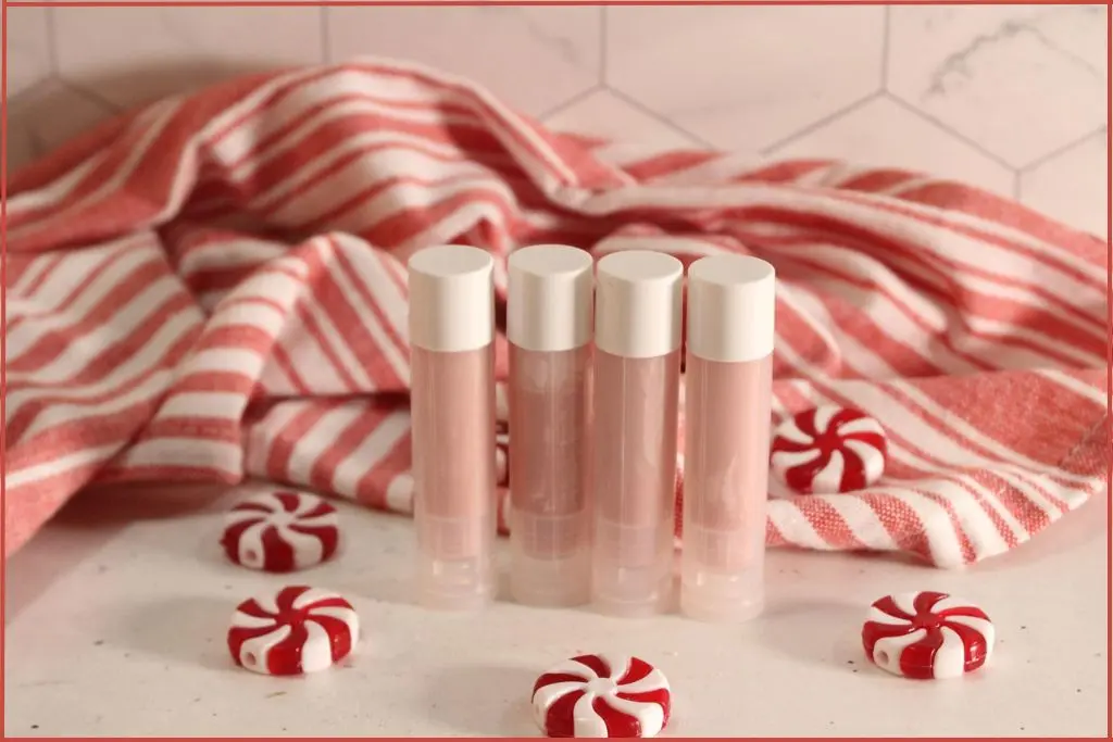 Four DIY candy cane lip balm tubes in front of a red‑and‑white striped towel, with peppermint candies scattered around.