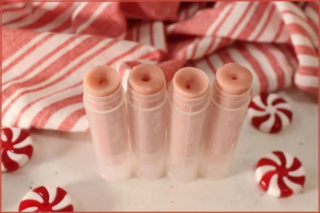 Four opened homemade candy cane lip balm tubes in front of a red‑and‑white striped towel, with peppermint candies scattered around.