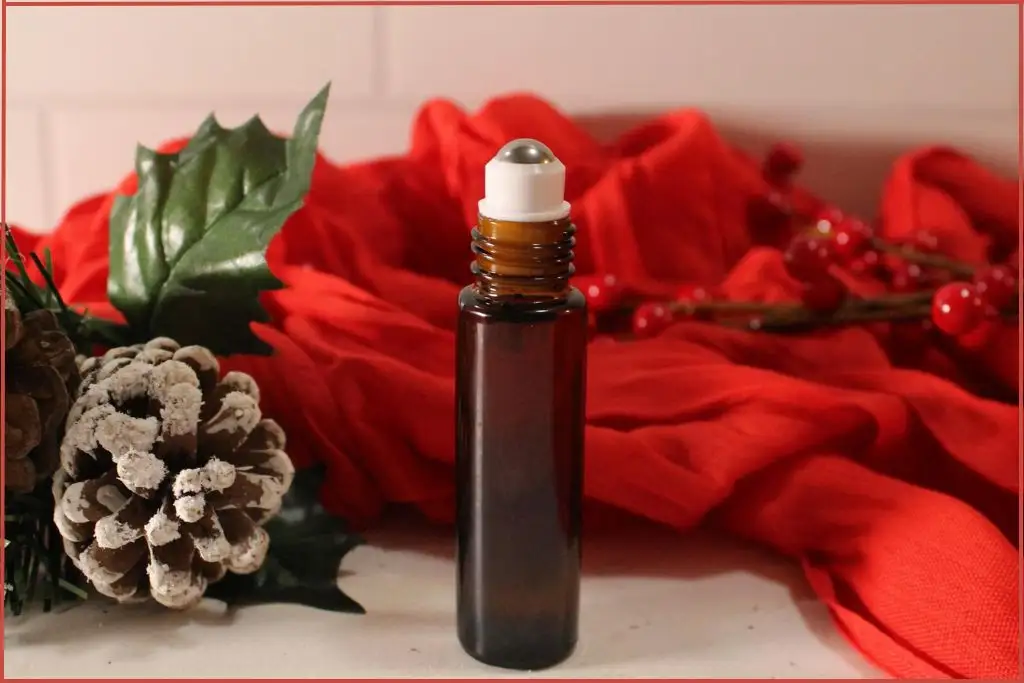 DIY Christmas roll-on perfume in a small amber bottle with roller ball, styled with red berries, frosted pinecones, and a red scarf backdrop