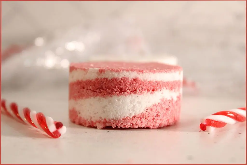 Side view of one round red-and-white layered candy cane bath bomb on a countertop, styled with mini candy canes with tips for success when making your own.