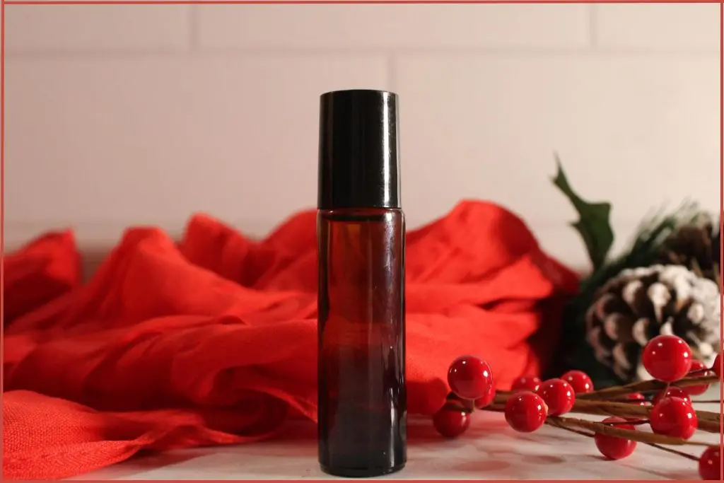 DIY Christmas roll-on perfume in a small amber bottle, styled with red berries, frosted pinecones, and a red scarf backdrop with tips for success