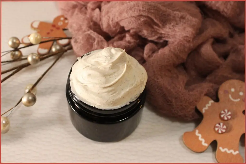 DIY gingerbread whipped body butter in a glass jar, creamy swirls on top, surrounded by 1 gingerbread man decorations, soft pink cloth, and holiday pearl stems on a white background.