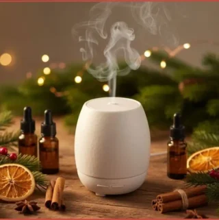White ceramic diffuser with traditional Christmas blends on wooden table surrounded by 3 essential oil bottles, cinnamon sticks, orange slices, and red berries