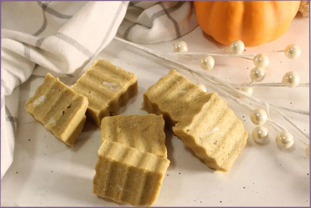 6 Rectangular homemade pumpkin spice sugar scrub bars with ridged tops arranged on a white surface, styled with a plaid kitchen towel, small orange pumpkin, and cozy pumpkin decor with instructions for how to make pumpkin spice sugar scrub bars.
