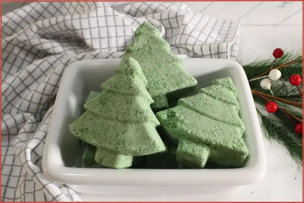 Christmas tree–shaped bath bombs stacked in a white dish, set on a checkered kitchen towel with pine greenery and red-and-white berry accents.