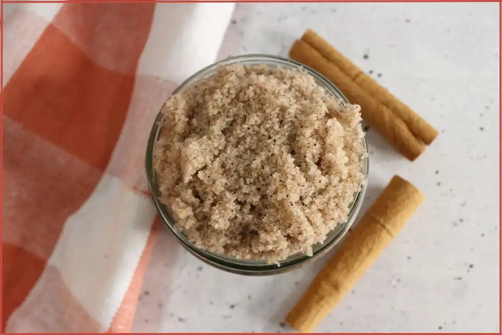 Brown sugar scrub in a glass jar surrounded by checked fabric. Two cinnamon sticks are on one side