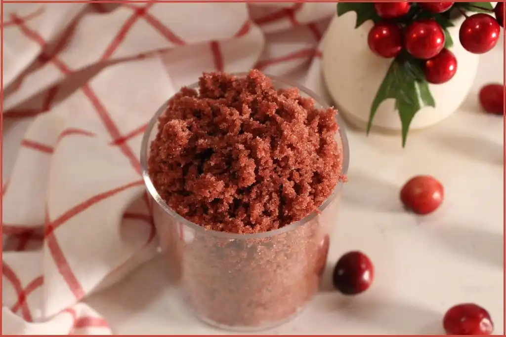 Rich red sugar scrub in a glass jar surrounded by cranberries and red and white checked fabric