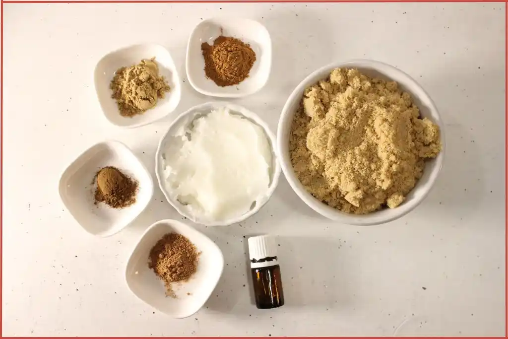 Gingerbread sugar scrub recipe ingredients - essential oil bottle and small white bowls with brown sugar, coconut oil, and powdered ginger, cinnamon, nutmeg, and clove.