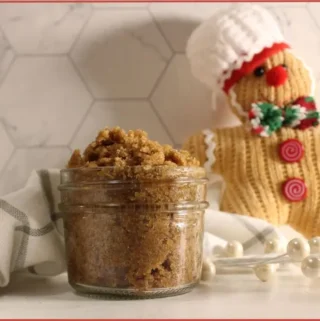 Small glass jar filled with brown gingerbread sugar scrub on a striped kitchen towel, with a knitted gingerbread man decoration wearing a chef hat in the background on a marble tile surface