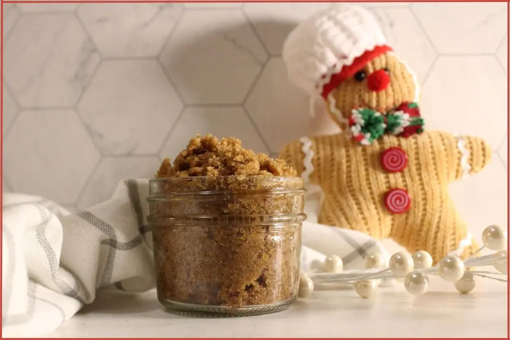 Small glass jar filled with brown gingerbread sugar scrub on a striped kitchen towel, with a knitted gingerbread man decoration wearing a chef hat in the background on a marble tile surface