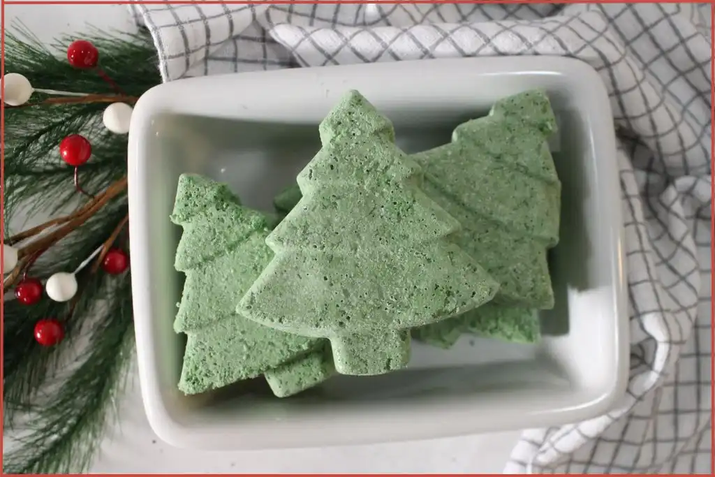 3 homemade Christmas tree–shaped bath bombs stacked in a white dish, set on a checkered kitchen towel with pine greenery and red-and-white berry accents.
