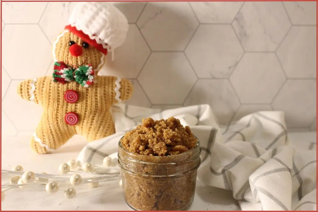 Small glass jar filled with brown gingerbread sugar scrub on a striped kitchen towel, with a knitted gingerbread man decoration wearing a chef hat in the background on a marble tile surface