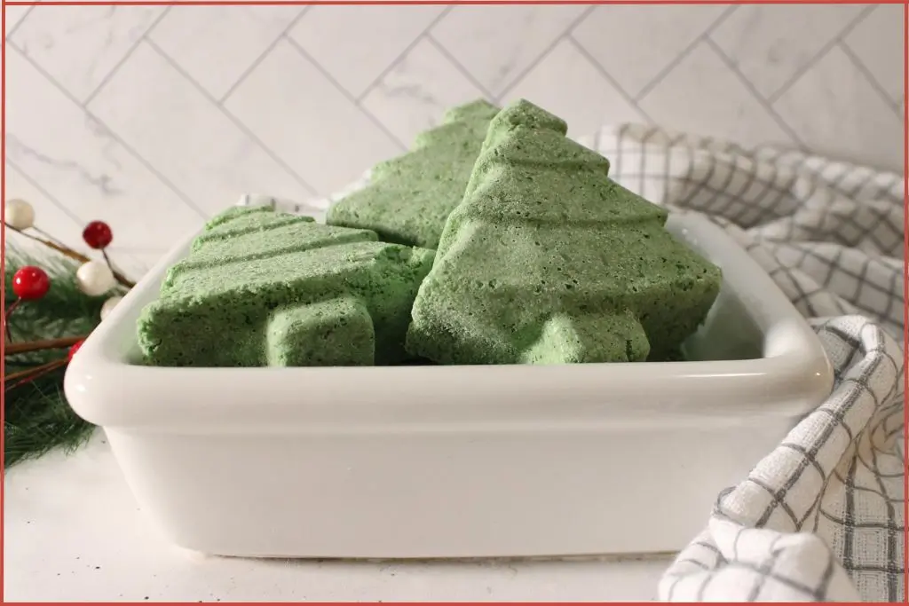 Christmas tree–shaped bath bombs stacked in a white dish, set on a checkered kitchen towel with pine greenery and red-and-white berry accents.