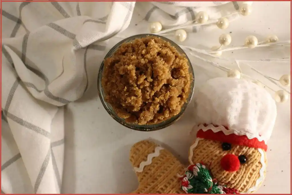 Top view of Small glass jar filled with brown gingerbread sugar scrub on a striped kitchen towel, with a knitted gingerbread man decoration wearing a chef hat in the background on a marble tile surface