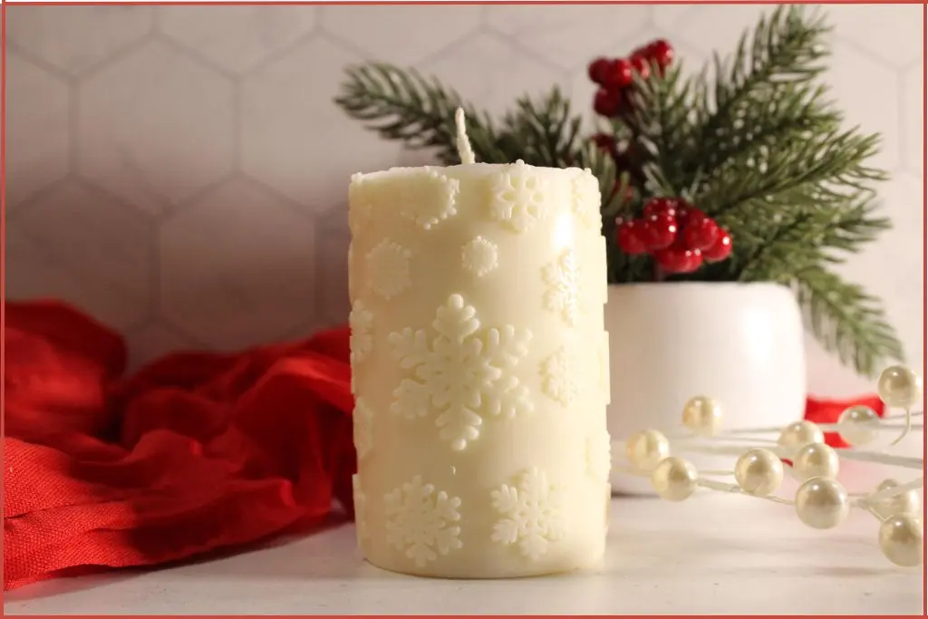 Homemade white snowflake pillar candle, styled on a white counter with a red cloth, winter greenery, and berries in the background.
