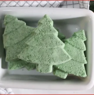 Homemade Christmas tree–shaped bath bombs stacked in a white dish, set on a checkered kitchen towel with pine greenery and red-and-white berry accents.