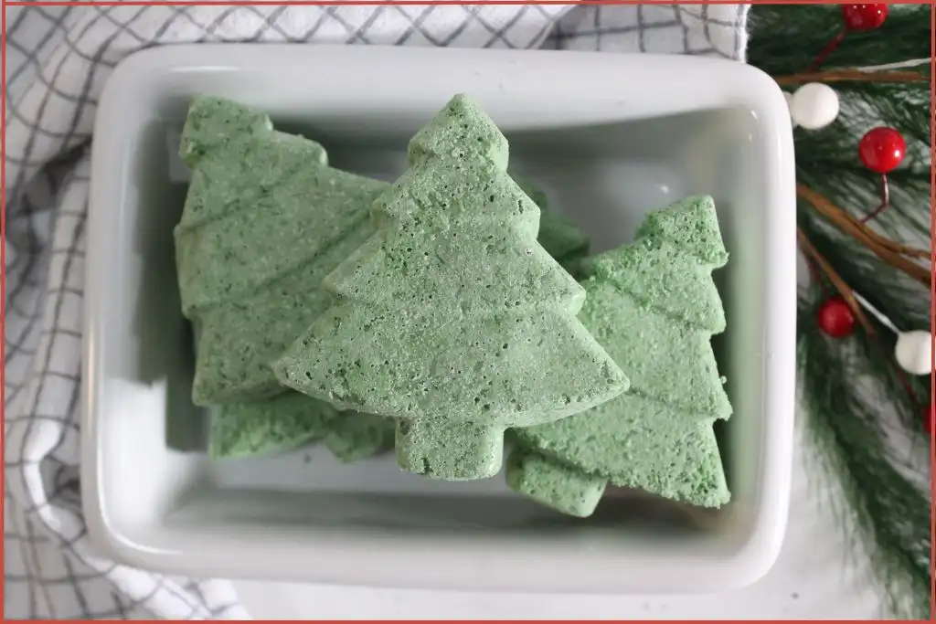 Homemade Christmas tree–shaped bath bombs stacked in a white dish, set on a checkered kitchen towel with pine greenery and red-and-white berry accents.