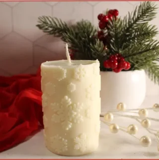 Homemade white snowflake candle made with essential oils, styled on a white counter with a red cloth, winter greenery, and berries in the background.