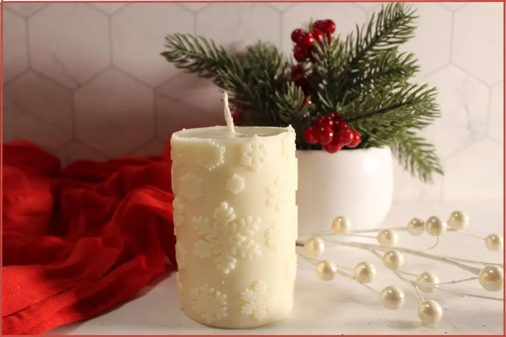 Homemade white snowflake candle made with essential oils, styled on a white counter with a red cloth, winter greenery, and berries in the background.