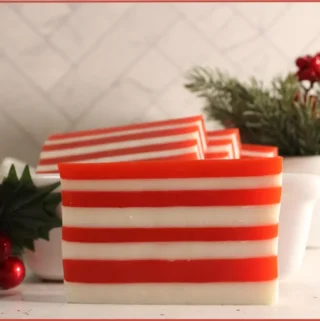 3 red and white striped candy cane soap bars on a wooden tray in a ceramic bowl with Christmas decorations at the side