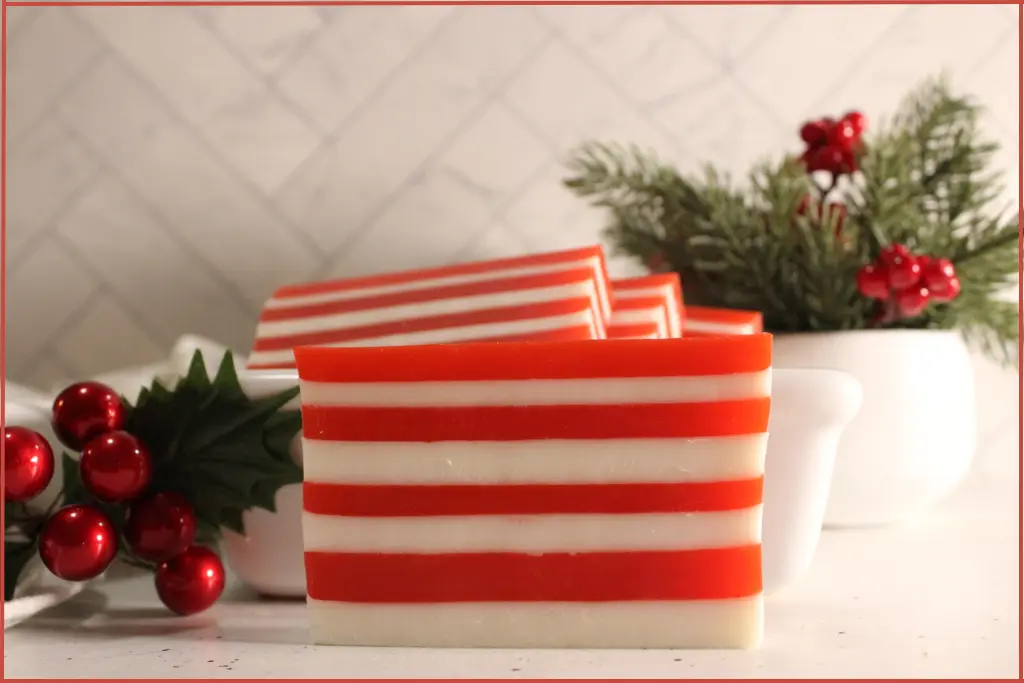 3 red and white striped candy cane soap bars on a wooden tray in a ceramic bowl with Christmas decorations at the side