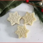 Three white snowflake-shaped sugar scrub bars on a marble surface with evergreen and ribbon, DIY recipe.