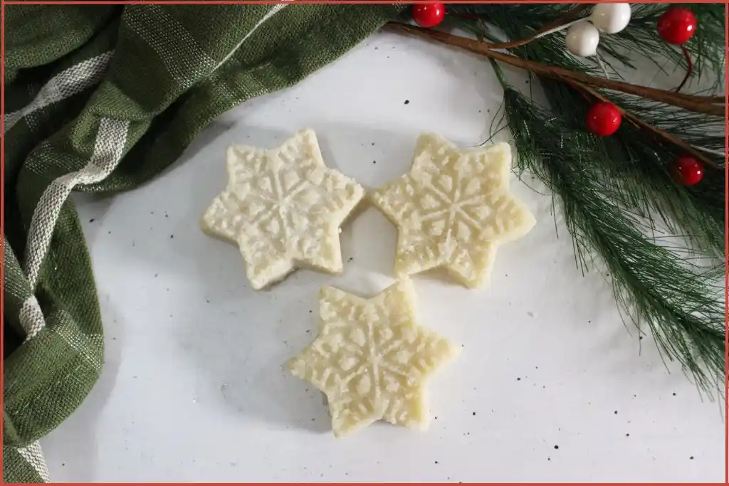 Three white snowflake-shaped sugar scrub bars on a marble surface with evergreen and ribbon, DIY recipe.