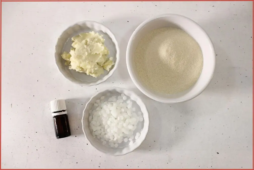 Ingredients for homemade snowflake sugar scrub bars - beeswax, shea butter and sugar in white bowls with an essential oil bottle at the side